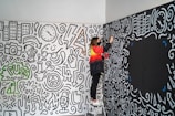 A person stands on a step ladder painting a wall covered with intricate, monochrome doodles. The illustrations feature various motifs such as buildings, clocks, lightning bolts, peace symbols, and music notes, along with some colored accents like a green tree with the word 'PUSH'. The person is wearing a red and black jacket and appears to be concentrating on adding more details to the artwork.