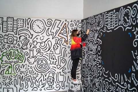 A person stands on a step ladder painting a wall covered with intricate, monochrome doodles. The illustrations feature various motifs such as buildings, clocks, lightning bolts, peace symbols, and music notes, along with some colored accents like a green tree with the word 'PUSH'. The person is wearing a red and black jacket and appears to be concentrating on adding more details to the artwork.