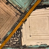 A detailed view of a textured ceiling featuring aged tin tiles with various colors and a diagonal wooden beam. The tiles exhibit a distressed look with peeling paint in shades of blue, off-white, and black, creating a vintage or rustic appearance.
