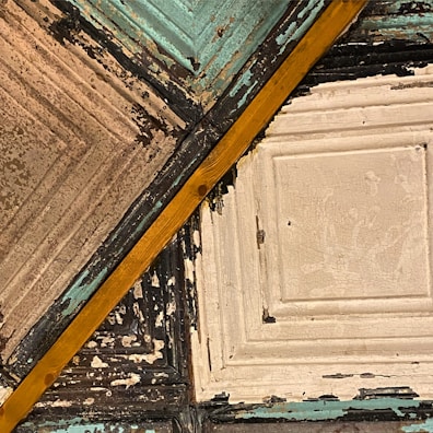A detailed view of a textured ceiling featuring aged tin tiles with various colors and a diagonal wooden beam. The tiles exhibit a distressed look with peeling paint in shades of blue, off-white, and black, creating a vintage or rustic appearance.