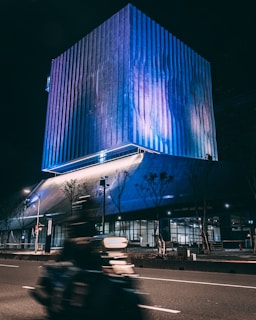 A large, modern building illuminated with vibrant blue and purple lights at night. Its design features vertical lines and a reflective surface that captures the light. A motorbike speeds along the street in front of the building, with motion blur conveying a sense of movement. Leafless trees line the sidewalk, and streetlights add to the ambient lighting.