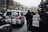 A street scene with a police vehicle parked alongside the road, accompanied by several pedestrians. The vehicle has markings indicating it is part of the local police force. There appears to be a person in uniform near the back of the vehicle, and another person holding a sign that is partially blurred. The environment is urban with buildings lining the street.