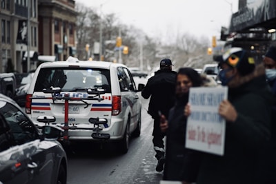 A street scene with a police vehicle parked alongside the road, accompanied by several pedestrians. The vehicle has markings indicating it is part of the local police force. There appears to be a person in uniform near the back of the vehicle, and another person holding a sign that is partially blurred. The environment is urban with buildings lining the street.