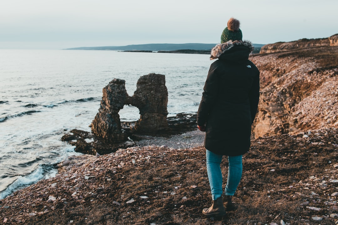 woman in black jacket and blue denim shorts standing on rocky shore during daytime