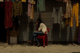 A person sits on a red plastic chair in front of a closed metal shutter, surrounded by hanging clothes of various styles and colors. The scene is dimly lit, creating a peaceful and contemplative atmosphere.