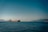 A large cargo ship sails through calm waters with a backdrop of distant mountains. The sky is clear, with a serene horizon separating the deep blue sea from the lighter shades of the sky.