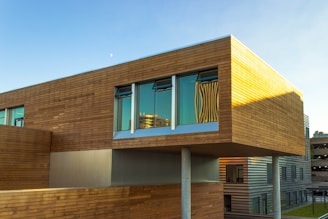 A modern architectural building featuring a design with horizontal wooden panels and large windows reflecting the sky. The structure appears elevated with visible support columns underneath, showcasing a blend of contemporary materials and finishes.