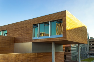 A modern architectural building featuring a design with horizontal wooden panels and large windows reflecting the sky. The structure appears elevated with visible support columns underneath, showcasing a blend of contemporary materials and finishes.