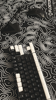 A mechanical keyboard with black keys and white keycaps on some keys rests on a patterned surface. A black computer mouse with holes in its shell and a black stand are positioned nearby. The surface features a black and white topographic design with contour lines, creating an abstract appearance.