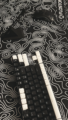 A mechanical keyboard with black keys and white keycaps on some keys rests on a patterned surface. A black computer mouse with holes in its shell and a black stand are positioned nearby. The surface features a black and white topographic design with contour lines, creating an abstract appearance.