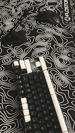A mechanical keyboard with black keys and white keycaps on some keys rests on a patterned surface. A black computer mouse with holes in its shell and a black stand are positioned nearby. The surface features a black and white topographic design with contour lines, creating an abstract appearance.