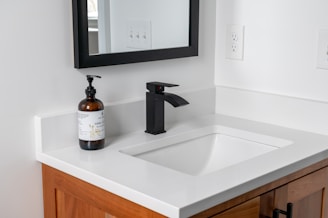 A modern bathroom sink with a sleek black faucet is set against a white countertop. A brown glass soap dispenser with a label is placed to the left of the faucet. The sink is integrated into a wooden vanity with visible grain patterns, and a black-framed mirror hangs above. An electrical outlet is visible on the wall next to the mirror.