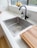 A modern kitchen setup featuring a stainless steel sink with a black faucet. On the countertop, a wooden cutting board is placed nearby, and a bottle of dish soap is next to the sink. A bowl of lemons sits on the windowsill above the sink, and the countertop is a light-colored stone material. Natural light enters through the window, creating a clean and bright atmosphere.