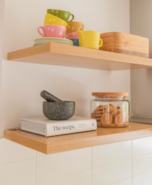 A neatly arranged kitchen shelf holding colorful stacked mugs and plates, a wooden container, and a second shelf below holding a mortar and pestle atop a cookbook titled 'The Recipe,' along with a glass jar filled with cookies.