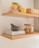A neatly arranged kitchen shelf holding colorful stacked mugs and plates, a wooden container, and a second shelf below holding a mortar and pestle atop a cookbook titled 'The Recipe,' along with a glass jar filled with cookies.