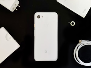 A flat lay of a white smartphone surrounded by its accessories on a dark background. The phone is centrally placed with a camera and fingerprint sensor visible on the back. Accompanying the phone are a charger, USB cable, SIM card removal tool, and packaging materials, all in a minimalistic and organized fashion.
