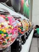 A row of gumball machines filled with colorful candies and chewing gum sits on a counter. The machines are transparent, allowing the vibrant assortment of pink, green, yellow, and white gum to be visible. A child in a green shirt is reaching towards one of the machines at the far end.