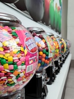 A row of colorful gumball machines filled with various types of candies. Each machine has a clear, round container showcasing vibrant, multicolored sweets including pink, yellow, green, white, and orange pieces. The machines are lined up on a white surface, and the perspective highlights the depth of the row, with some machines slightly out of focus in the background. Bright packaging labels are visible, displaying the Dubble Bubble brand and images of gum.