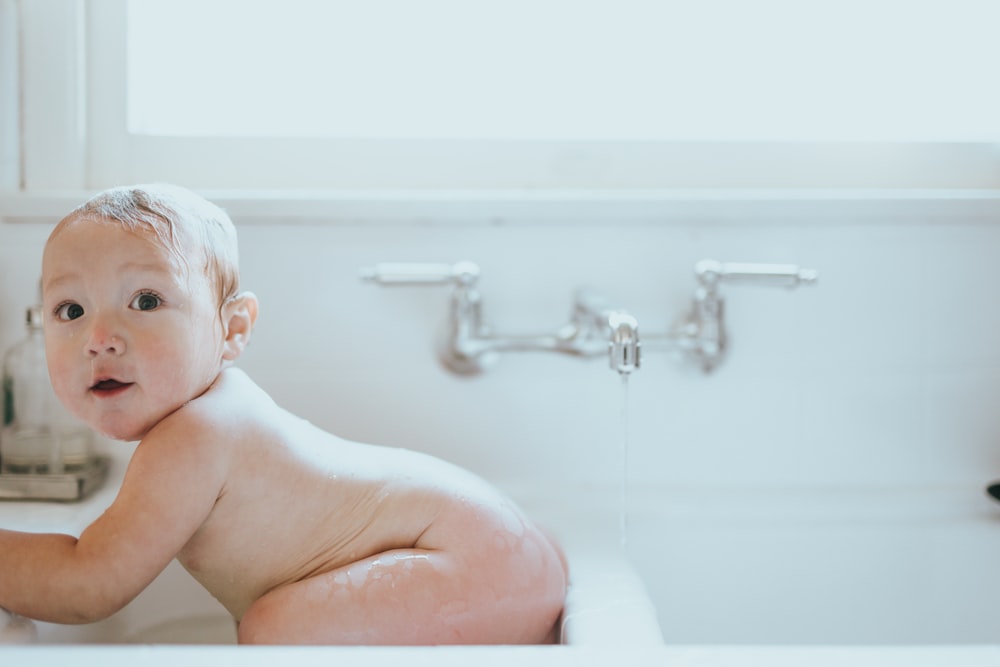 naked baby lying on white ceramic bathtub