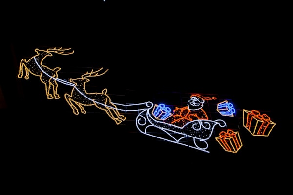A neon-style light display features a depiction of Santa Claus riding in a sleigh pulled by two reindeer. The sleigh is filled with colorful wrapped presents, highlighting festive red, blue, and yellow colors against a dark background.