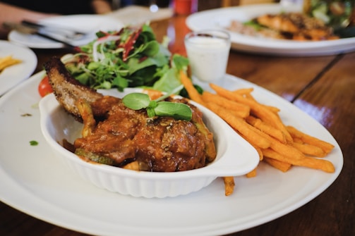 A well-plated dish featuring a portion of meat with a rich sauce, accompanied by a side of sweet potato fries and a fresh green salad. A small cup of creamy dipping sauce sits beside the fries, all arranged on a white oval plate on a wooden table.