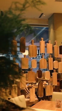 A series of wooden cutting boards are suspended in front of a cozy restaurant interior, soft lighting illuminating the warm wooden tones. Behind the boards, seating areas with plush chairs and tables can be seen.