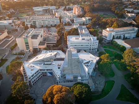 An aerial view of an urban area featuring multiple buildings of varied architectural styles, surrounded by trees and green spaces. The structures are dense with a mix of flat and angled rooftops, suggesting a campus or institutional setting. A sports field is visible in the background, indicating recreational facilities nearby.