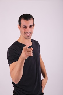 A man with short black hair and facial hair is smiling and pointing directly at the camera. He is wearing a black t-shirt and has one hand on his hip.