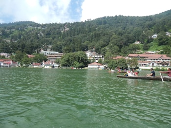 The image captures a serene lake surrounded by lush green hills and a line of charming buildings along the shore. Several boats with people enjoying rides are present on the water, creating a sense of leisurely activity.