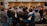 A group of people standing in a circle, with their arms around each other, suggesting a sense of community or solidarity. In the background, there are individuals facing the group, possibly leading or speaking to them. The setting appears to be indoors, with a focus on interaction and connection among the participants.