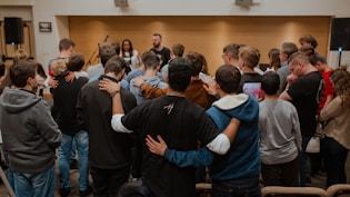 A group of people standing in a circle, with their arms around each other, suggesting a sense of community or solidarity. In the background, there are individuals facing the group, possibly leading or speaking to them. The setting appears to be indoors, with a focus on interaction and connection among the participants.