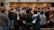 A group of people standing in a circle, with their arms around each other, suggesting a sense of community or solidarity. In the background, there are individuals facing the group, possibly leading or speaking to them. The setting appears to be indoors, with a focus on interaction and connection among the participants.