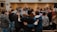 A group of people standing in a circle, with their arms around each other, suggesting a sense of community or solidarity. In the background, there are individuals facing the group, possibly leading or speaking to them. The setting appears to be indoors, with a focus on interaction and connection among the participants.