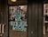 Large, bold blue and yellow letters on a glass door read 'Buy Local or Bye-Bye Local', reflecting a street scene with buildings and pedestrians in the background.