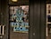 Large, bold blue and yellow letters on a glass door read 'Buy Local or Bye-Bye Local', reflecting a street scene with buildings and pedestrians in the background.