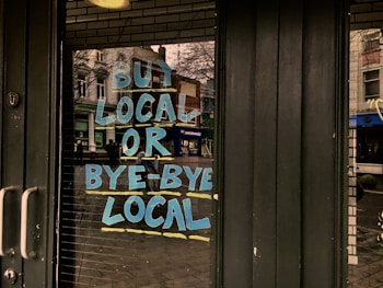 Large, bold blue and yellow letters on a glass door read 'Buy Local or Bye-Bye Local', reflecting a street scene with buildings and pedestrians in the background.