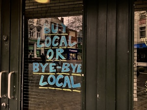 Large, bold blue and yellow letters on a glass door read 'Buy Local or Bye-Bye Local', reflecting a street scene with buildings and pedestrians in the background.