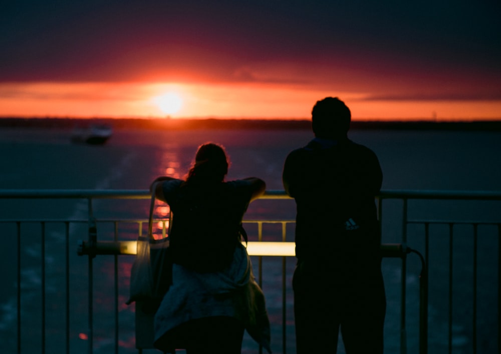silhouette of man and woman standing beside railings during sunset