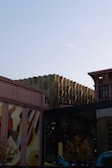 A modern urban scene with architectural structures featuring a mix of geometric patterns and reflective glass surfaces. The structures include a uniquely textured golden building, a facade with colorful abstract art, and mirrored windows reflecting the environment. Construction machinery is visible at the bottom, suggesting ongoing development.