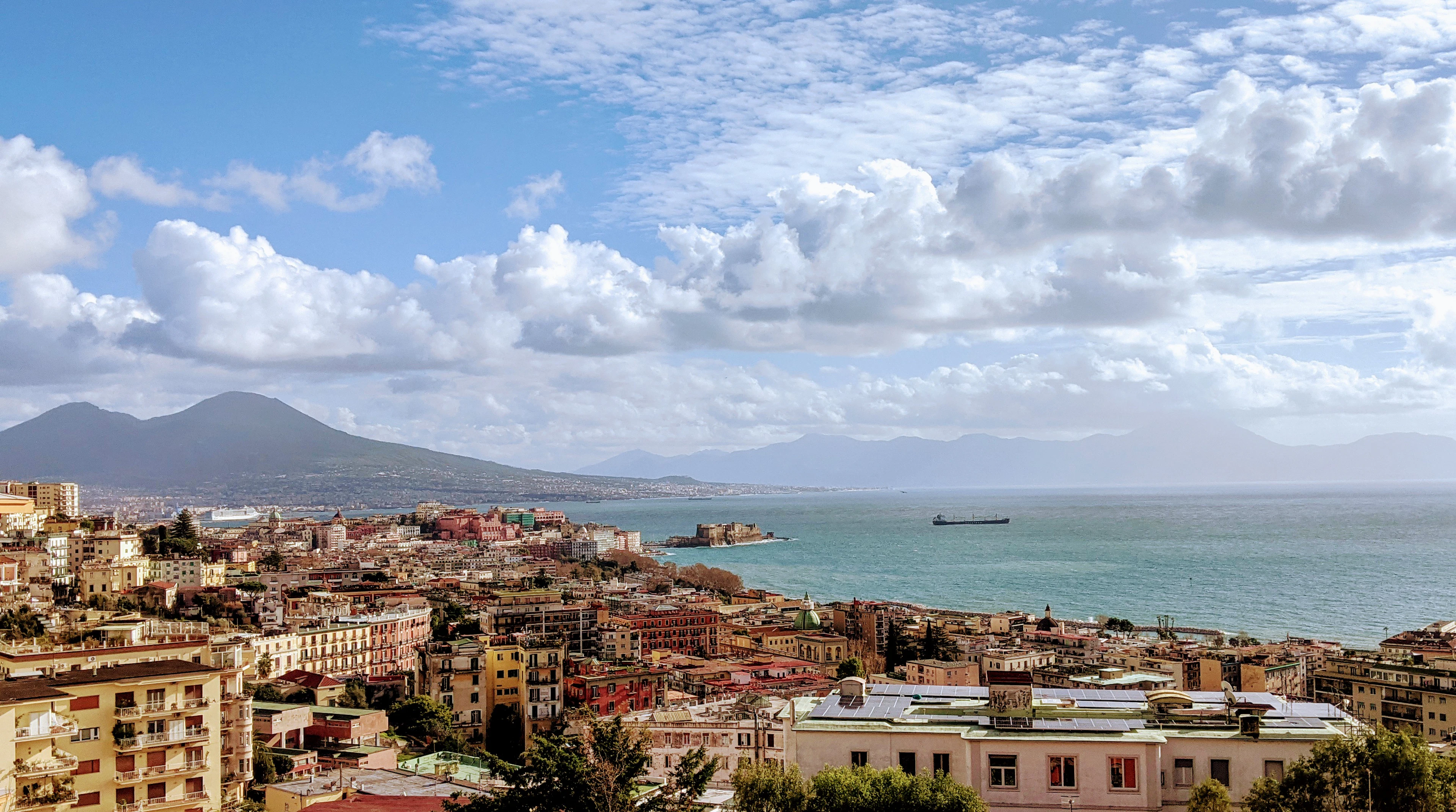 The Bay of Naples seen from above, pale apartment blocks running down to the water with the cone of Vesuvius on the horizon