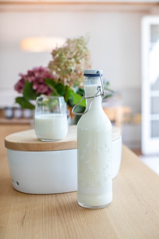 A glass bottle filled with milk and sealed with a flip-top lid is placed on a wooden table in the foreground. Directly behind it is a glass half-filled with milk, positioned on a white ceramic object with a wooden lid. In the background, a centerpiece with a mix of drying and fresh flowers, mainly in shades of green and purple, is visible. The setting appears to be a cozy, well-lit room with blurred details like furniture and a lamp visible.