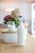 A glass bottle filled with milk and sealed with a flip-top lid is placed on a wooden table in the foreground. Directly behind it is a glass half-filled with milk, positioned on a white ceramic object with a wooden lid. In the background, a centerpiece with a mix of drying and fresh flowers, mainly in shades of green and purple, is visible. The setting appears to be a cozy, well-lit room with blurred details like furniture and a lamp visible.
