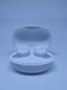 A pair of white wireless earbuds in an open charging case, set against a light blue background. The earbuds are clearly marked with 'R' and 'L' for right and left orientation, and the case displays certification symbols.