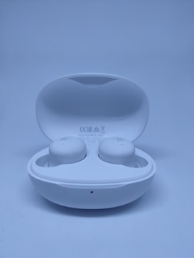 A pair of white wireless earbuds in an open charging case, set against a light blue background. The earbuds are clearly marked with 'R' and 'L' for right and left orientation, and the case displays certification symbols.