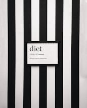 A framed definition of the word 'diet' displayed on a wall with vertical black and white stripes. The definition humorously states 'always starts tomorrow' beneath a phonetic spelling.