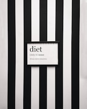 A framed definition of the word 'diet' displayed on a wall with vertical black and white stripes. The definition humorously states 'always starts tomorrow' beneath a phonetic spelling.
