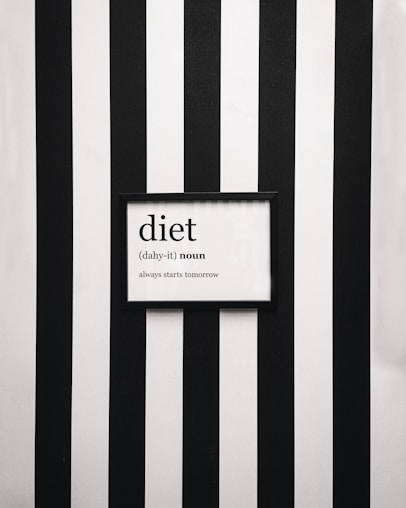 A framed definition of the word 'diet' displayed on a wall with vertical black and white stripes. The definition humorously states 'always starts tomorrow' beneath a phonetic spelling.