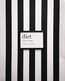 A framed definition of the word 'diet' displayed on a wall with vertical black and white stripes. The definition humorously states 'always starts tomorrow' beneath a phonetic spelling.