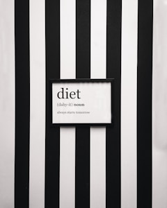 A framed definition of the word 'diet' displayed on a wall with vertical black and white stripes. The definition humorously states 'always starts tomorrow' beneath a phonetic spelling.