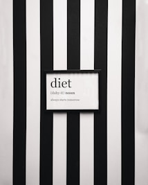 A framed definition of the word 'diet' displayed on a wall with vertical black and white stripes. The definition humorously states 'always starts tomorrow' beneath a phonetic spelling.
